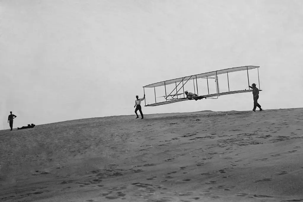 pioneers-of-aviation-closing Image of Wilbur Wright (piloting the plane) and his brother Orville, holding onto the plane (L), at Kitty Hawk, NC.